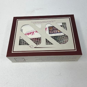May include: A rectangular craft kit box with a maroon border and a cream-colored interior. The box has a die-cut window revealing the contents, including a needle and thread. Text on the box reads "Peek through our window and see the fine quality materials packed in this kit."
