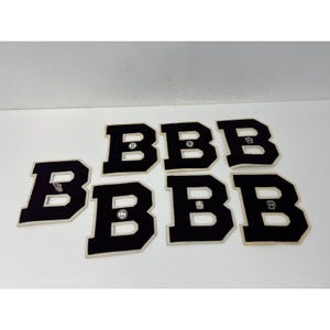 May include: Six black felt letter "B" patches with white trim and a small emblem in the center. The patches are arranged on a white surface. The letters are in a bold, block style.