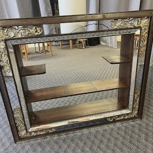 May include: A rectangular wall mirror with a dark brown frame and gold filigree accents. The mirror has three wooden shelves and a mirrored interior, creating a display case. The frame is approximately 30 inches wide.