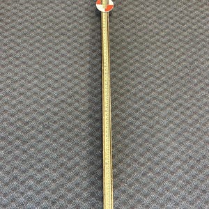 May include: A vintage wooden folding ruler, featuring a white and red circular design at the top. The ruler is marked with inch measurements. The ruler is a light brown color and is lying on a dark gray patterned surface.