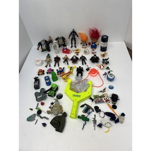 May include: A collection of vintage toys, including action figures, trolls with colorful hair, toy cars, and a neon green slingshot. Various small plastic figures and accessories are scattered across a white surface.