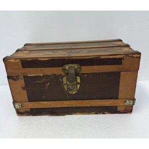 May include: A rectangular storage trunk featuring a two-tone brown and tan exterior with horizontal wooden slats. The chest includes a central metal latch mechanism with a weathered finish and reinforced metal corner protectors on the edges.