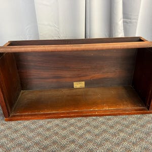 May include: A rectangular wooden storage box with a handle. The box is made of dark brown wood with a lighter brown handle. A small rectangular label is affixed to the front of the box. The box is empty.