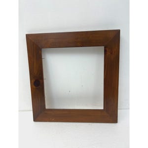 May include: A square wooden picture frame with a medium brown stained finish. The frame features a wide, flat border with visible wood grain and a prominent dark knot on the left side. The inner edges are angled inward toward the center.
