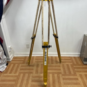 May include: A yellow and wood surveyor's tripod with the brand name "BERGER" printed vertically on the center leg. The tripod has three legs with metal tips and black straps. The legs are made of wood and painted yellow.