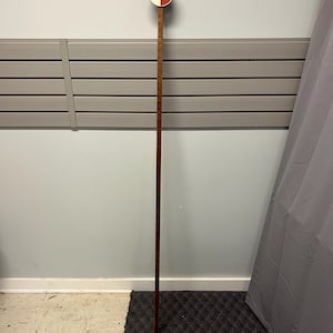 May include: A vintage wooden measuring stick with a circular red, white, and brown top. The stick is long and slender, with measurement markings. The top has the number 1. The stick is hanging on a wall.
