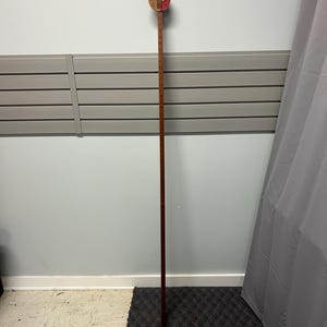 May include: A tall, wooden measuring stick with a circular top. The top is red and tan with a circular design. The stick is brown and has measurement markings. The stick is standing upright against a gray wall.