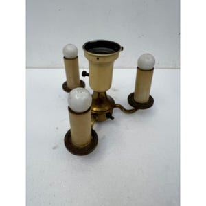 May include: A brass light fixture component featuring a central socket housing surrounded by three vertical candle-style sleeves, each topped with a small white spherical bulb. The metal base has a metallic gold finish with dark brown accents.