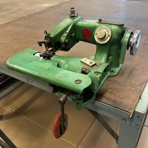 May include: A vintage green sewing machine with a red and white logo and a silver hand wheel. The machine has a red wheel attached to the side and is set on a brown table.