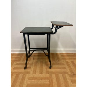 May include: A vintage school desk featuring a dark rectangular writing surface supported by a black metal frame with four legs on casters. An attached smaller rectangular side shelf extends from the right side, supported by a black metal bracket.