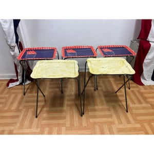 May include: A set of five folding TV tray tables with thin metal legs. Three trays feature a dark blue surface with a red and white polka dot border and a pocket graphic. Two trays have a textured, light yellow marbled surface finish.