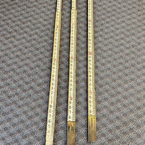 May include: Three vintage wooden folding rulers with black and red markings. Each ruler is numbered in inches, with a brass-colored metal end piece. The rulers are laid out on a textured gray surface.