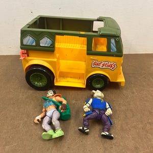 May include: A yellow and green toy van with the text "Foot Stinks" on the side, accompanied by two action figures. The van has black wheels and a green roof. One figure wears a blue top and purple pants, the other wears a green bag.