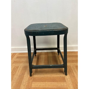 May include: A vintage, teal-colored metal stool with a square seat and four legs. The seat has small holes and a slightly worn appearance. The legs are connected by horizontal bars for added stability. The stool is placed on a wooden floor.