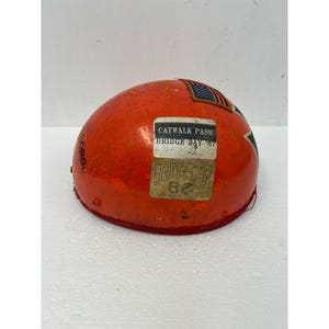 Puede incluir: Un casco de seguridad de color naranja brillante con un pequeño parche de la bandera estadounidense y dos pegatinas rectangulares. Una pegatina dice CATWALK PASS BRIDGE DAY '87 y la otra BRIDGE DAY 86. El lateral tiene la palabra GORGL en tinta negra.