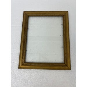 May include: A rectangular picture frame with a metallic gold finish and a textured, carved border pattern. The frame is empty, showing the inner edges with small metal pins used for securing a backing or glass, set against a plain white surface.