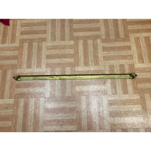 May include: A long, narrow, horizontal brass rod with a smooth, metallic finish. The rod features small, rounded mounting brackets at both ends, designed for attachment. The surface of the brass shows subtle variations in tone and texture.