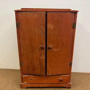 May include: A small, antique wooden cabinet painted in a faded reddish-orange hue. The cabinet has two doors with round wooden knobs and a small drawer at the bottom. The doors are slightly ajar, revealing the interior. Brass hinges and hardware are visible.