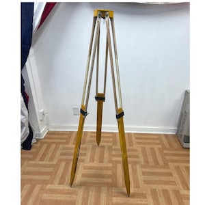May include: A vintage wooden tripod with yellow painted legs and natural wood supports. The tripod has a pointed base and is secured with black straps. The tripod is likely used for surveying or photography.