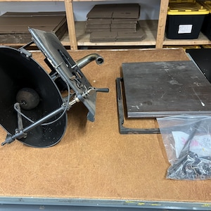 May include: An antique black gramophone horn and mechanical components are displayed on a wooden surface. A rectangular metal plate and a bag of hardware are also present. The items appear to be vintage and in a workshop setting.