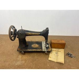 Op de afbeelding: Een vintage zwarte Singer naaimachine met gouden bloemmotieven, een handwiel en een metalen steekplaat. Inbegrepen zijn een rechthoekige houten opbergdoos, een papieren handleiding met de tekst No. 27-4 en diverse kleine metalen naaigereedschappen.