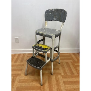 May include: A vintage metal step stool chair with a gray seat and backrest. The frame is white with gray accents. It features two retractable steps with worn black and yellow paint surfaces, showing signs of heavy use and chipped paint throughout.