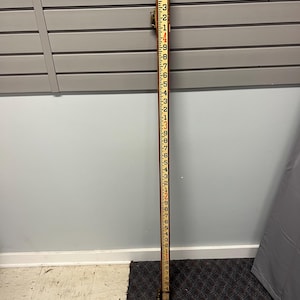 May include: A vintage wooden measuring stick with black and red markings. The stick is marked in feet and inches, with numbers and lines indicating measurements. The wood has a worn appearance, suggesting age and use. The stick is used for measuring height or depth.