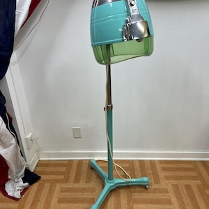 May include: Vintage teal and chrome hair dryer on a stand. The hood is a rounded shape with a clear plastic visor. The stand is teal with a three-legged base on wheels. A power cord hangs down the stand.
