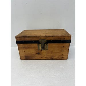 May include: A rectangular wooden storage chest with a natural light brown finish. The box features a horizontal black metal band across the front, a central brass-colored latch mechanism, and visible small nail heads along the edges of the wood.