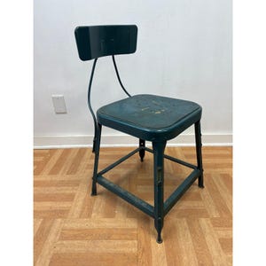 May include: Vintage teal metal chair with a square seat and small backrest. The chair has a sturdy, industrial design with a triangular base and a curved back support. The seat has small holes and shows signs of wear.