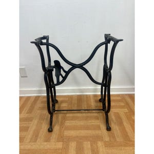 May include: A black cast iron sewing machine base featuring an ornate, curved frame with four legs, a central cross-brace, and a small decorative figure integrated into the side structure. The metal frame is open and designed for a tabletop.