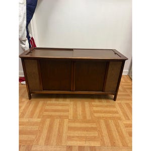 May include: A rectangular wooden media console featuring a dark brown finish, a flat top surface with sliding panels, and two vertical side sections covered in a textured, light-colored woven fabric mesh. The unit stands on four short legs.