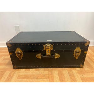 May include: A rectangular black trunk featuring weathered brass-toned metal hardware, including corner reinforcements, two side latches, and a central locking mechanism with a handle. The exterior surface displays visible scuffs and signs of wear.