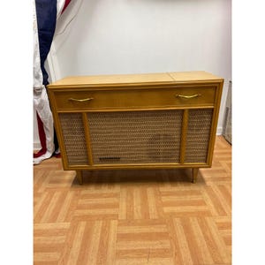 May include: A rectangular wooden console cabinet with a light brown finish, featuring two gold-toned metal handles on the top drawer section and a woven cane speaker grille covering the lower front panel, supported by four short tapered legs.