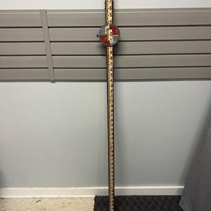 May include: A vintage wooden measuring stick with black and red markings, featuring numbers from 1 to 9. A circular, metal gauge is attached at the center. The stick is approximately 9 feet tall.