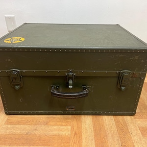 May include: Olive green metal trunk with a black leather handle and metal latches. The trunk has a yellow star emblem on the top left corner. The trunk is riveted and has a rectangular shape.