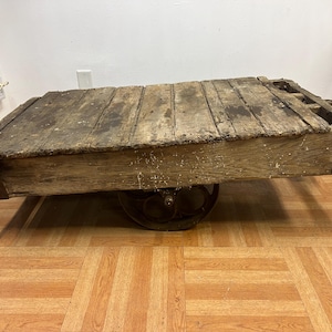 May include: An antique wooden cart with a weathered appearance. The cart features a rectangular platform made of wooden planks and is supported by two metal wheels. The wood has a distressed look, with visible grain and signs of age.