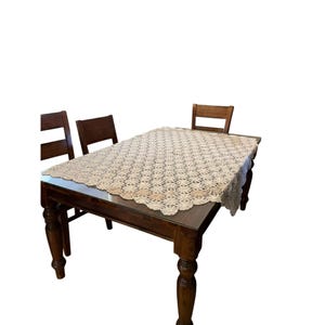 May include: A dark brown wooden dining table with a beige lace tablecloth. The table has four turned legs and is surrounded by three matching wooden chairs. The tablecloth has a floral pattern.
