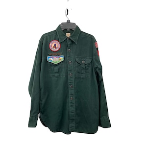 May include: Dark green button-down shirt with two chest pockets and patches. Patches include a circular emblem with text and a landscape scene. The shirt is hanging on a black hanger against a white background.