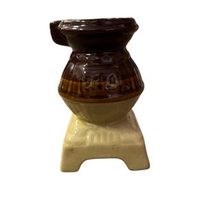 May include: A small, ceramic decorative object with a dark brown top, a lighter brown midsection, and a cream-colored base. The object has a handle on the side and a textured pattern on the midsection. The base is rectangular with cut-out sections.