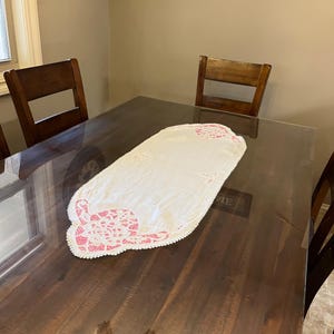 May include: A white table runner with pink floral embroidery and a scalloped edge rests on a dark wooden table. The runner is rectangular with decorative detailing at each end. The table is surrounded by wooden chairs.