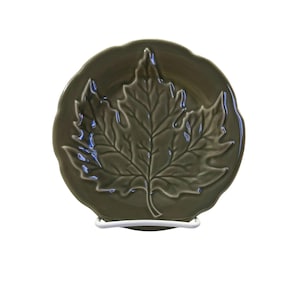 May include: A ceramic plate with a raised leaf design in a muted green color. The plate has a scalloped edge and is displayed on a white metal stand. The leaf's veins are detailed, adding texture to the plate's surface.