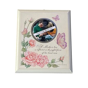 May include: A rectangular white picture frame featuring a circular center cutout. The frame is decorated with pink roses, green leaves, and butterflies. The text reads: A Mother's love is reflected in the joyful faces of her loved ones. Insert Your photo here.