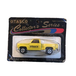 May include: A yellow die-cast metal OTASCO Collector's Series pickup truck toy in its original packaging. The truck has black wheels and the OTASCO logo on the side. The packaging is black with the text "Collector's Series".