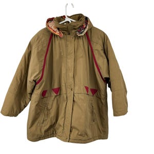 May include: A tan hooded jacket with red trim and accents. The jacket has a patterned hood lining and two front pockets. The jacket is hanging on a white hanger.