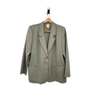 Puede incluir: Un blazer verde salvia con cierre de un solo botón y dos bolsillos delanteros. El blazer está colgado de una percha de madera sobre un fondo blanco. El blazer tiene un diseño clásico y entallado.