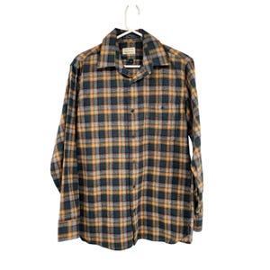 May include: A long-sleeved, button-down flannel shirt in a plaid pattern of green, gold, and gray. The shirt has a collared neckline, a chest pocket, and is hanging on a white hanger.