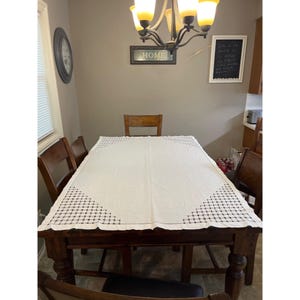 May include: A rectangular white tablecloth featuring a decorative black grid pattern at each of the four corners. The tablecloth is draped over a dark brown wooden dining table surrounded by matching wooden chairs in a room with a chandelier.