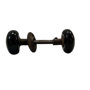 May include: A vintage door knob set with two glossy black oval knobs connected by a rusty metal rod and a circular plate. The hardware is designed for a door and is set against a white background.