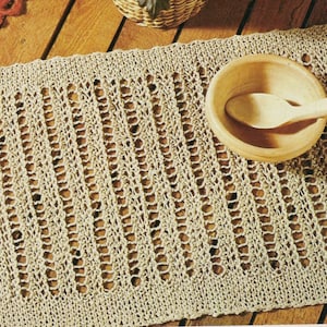 May include: A beige crocheted placemat with a woven pattern. The placemat is on a wooden surface with a wooden bowl and spoon on top.